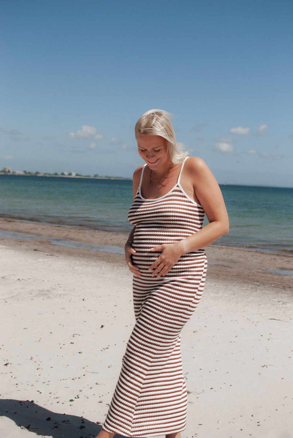 Am Strand: Die Schwangere schaut lächelnd auf ihren Babybauch, dahinter das Meer